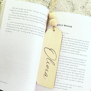 May include: A wooden bookmark with a light brown finish and a white string attached to the top. The bookmark has the name "Olivia" engraved in cursive on the front. The bookmark is lying on a white page with text.