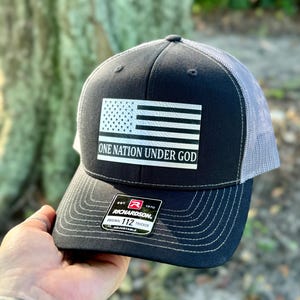 May include: A black and grey trucker hat with a white American flag and the text "One Nation Under God". The hat has a black and white Richardson label with the text "Original 112 Trucker Adjustable".