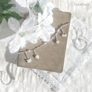 Puede incluir: Un delicado collar plateado con colgantes en forma de mariposas, corazones y estrellas, sobre un cuaderno marrón estampado. El collar está adornado con pequeñas cuentas. Encaje blanco y flores en el fondo.