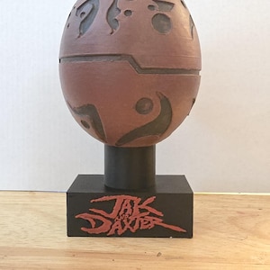 May include: A brown egg-shaped object with dark recessed markings and a horizontal seam, mounted on a black cylindrical post and a rectangular black base featuring the red text Jak and Daxter printed on the front face of the base.