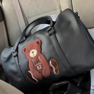 Puede incluir: Bolso de lona negro con un motivo de oso de peluche marrón. El oso de peluche lleva una camiseta con el número 82. El bolso tiene dos asas y una correa para el hombro. El bolso es de material liso.