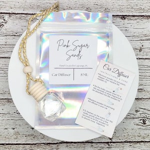 May include: A clear glass bottle with a wooden lid and a string for hanging. The bottle is filled with a clear liquid and is labeled "Car Diffuser" and "8 ML". The bottle is on a white plate with a holographic iridescent bag behind it. The bag is labeled "Pink Sugar Sands" and "Hand Created in Lagrange, IN". A card with instructions for using the car diffuser is also visible.
