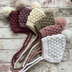May include: A selection of knitted baby bonnets in shades of burgundy, mauve, cream, olive green, pink, and white. Each bonnet has a textured design, ties, and a fluffy pom-pom. The bonnets are displayed against a brick background.