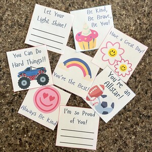 Può includere: Una collezione di biglietti quadrati con messaggi positivi e illustrazioni. I biglietti presentano frasi come "Lascia che la tua luce risplenda!" e "Sei il migliore!" con immagini di un camion mostro e un arcobaleno.