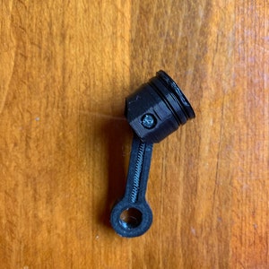 May include: A black plastic model of a piston and connecting rod. The piston is cylindrical with a small hole in the side. The connecting rod is a long, thin piece of plastic with a round end.