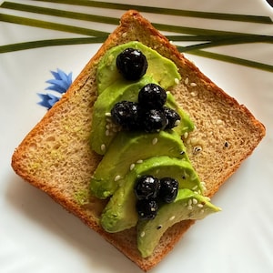 May include: A square slice of toasted brown bread topped with thin, green avocado slices arranged in a triangular shape. The avocado is garnished with dark purple blueberries and a light sprinkling of small white sesame seeds.