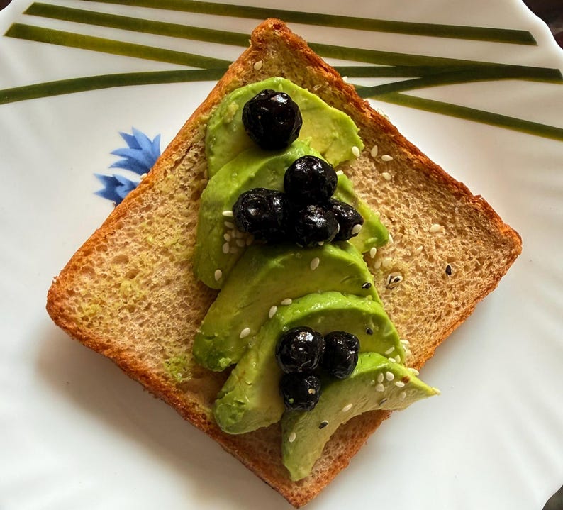 May include: A square slice of toasted brown bread topped with thin, green avocado slices arranged in a triangular shape. The avocado is garnished with dark purple blueberries and a light sprinkling of small white sesame seeds.