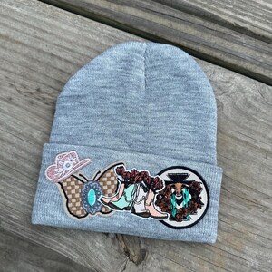 May include: A gray beanie with a variety of colorful patches, including a butterfly, cowboy boots, and a cactus with a bull's head.