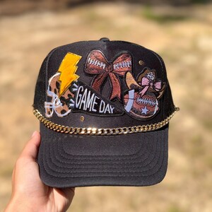 May include: Black trucker hat with a gold chain and football-themed patches. Patches include a football helmet, a bow, a football with a cowboy hat, and a lightning bolt. The text "GAME DAY" is also featured on the hat.