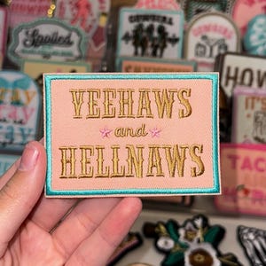 May include: A pink and turquoise embroidered patch with the text "Yeehaws and Hellnaws" in gold lettering.