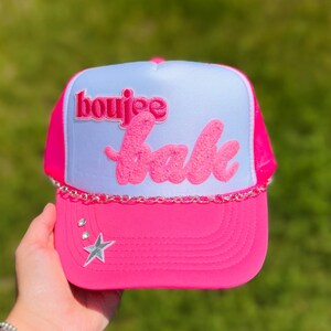 May include: A pink trucker hat with a white front panel. The hat features the text "boujee babe" in pink, a silver chain, and rhinestone accents. A silver star is also on the front.