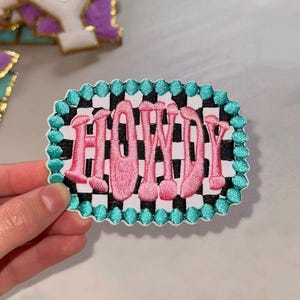 May include: Pink embroidered patch with the word "HOWDY" in a checkered black and white pattern with a turquoise border.
