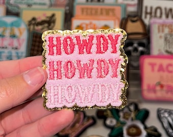 May include: A pink embroidered patch with the word "HOWDY" repeated three times in a stacked design. The patch has a gold glitter border and is held in a hand. Other patches are visible in the background.