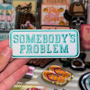 May include: White embroidered patch with teal blue lettering that reads "Somebody's Problem".