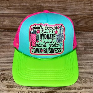 May include: A bright turquoise and neon green trucker hat with a pink mesh back. The front features a patch with the text "don't forget to HYDRATE and mind your OWN BUSINESS" alongside an illustration of a pink tumbler and a skeleton hand.