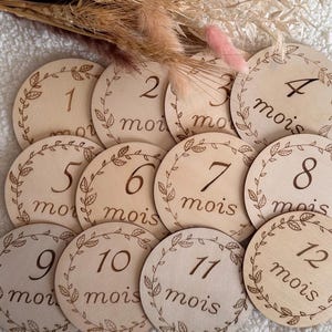 Op de afbeelding: Een set van twaalf ronde houten schijven, elk gegraveerd met een cijfer van 1 tot 12 en het woord mois, omringd door een delicate bladrand. De schijven zijn in rijen gerangschikt, vergezeld door een boeketje van gedroogde tarwe en roze gras.