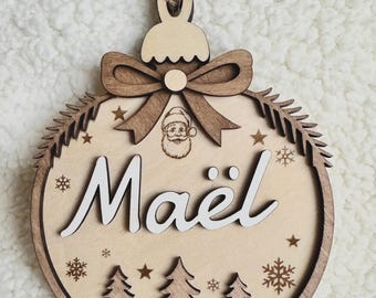 Puede incluir: Un adorno navideño redondo de madera con el nombre Maël en letras blancas. El diseño incluye un lazo cortado a láser, una pequeña cara de Papá Noel, ramas de pino, estrellas y copos de nieve, con un cordel de yute para colgar.