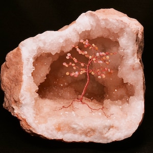 May include: A geode with a reddish-brown exterior and a sparkling, crystal-lined interior. A miniature tree sculpture with a red wire trunk and branches with pink beads is displayed inside the geode.
