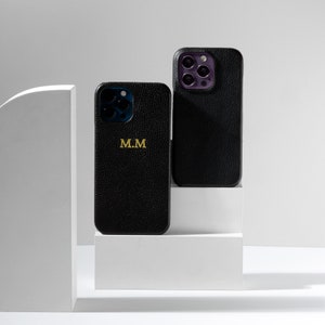 May include: Two black leather phone cases with a gold monogram. The case on the left has the initials "M.M." The case on the right has a camera lens cutout.