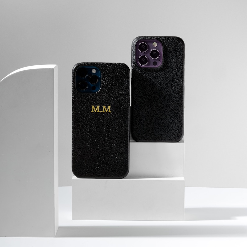 May include: Two black leather phone cases with a gold monogram. The case on the left has the initials "M.M." The case on the right has a camera lens cutout.