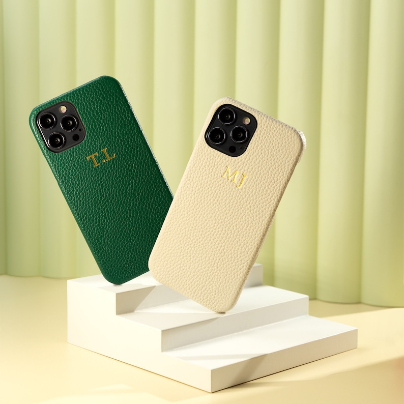 May include: Two personalized phone cases, one green and one cream, with initials "T.L." and "M.J."  The cases are made of textured leather and are displayed on a white tiered platform.