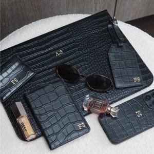 May include: A collection of black faux-leather accessories with a crocodile skin texture. Items include a clutch, wallet, phone case, luggage tag, and sunglasses. Gold initials are embossed on several items. A bottle of perfume and a tube of makeup are also present.