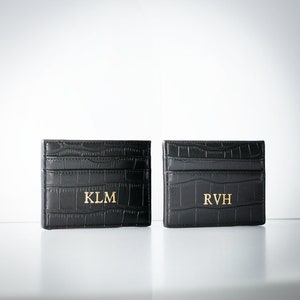 K&ouml;nnte beinhalten: Zwei schwarze Lederkartenhalter mit einem Krokodilpr&auml;gemuster. Die Kartenhalter haben goldene Beschriftungen mit den Initialen "KLM" und "RVH".