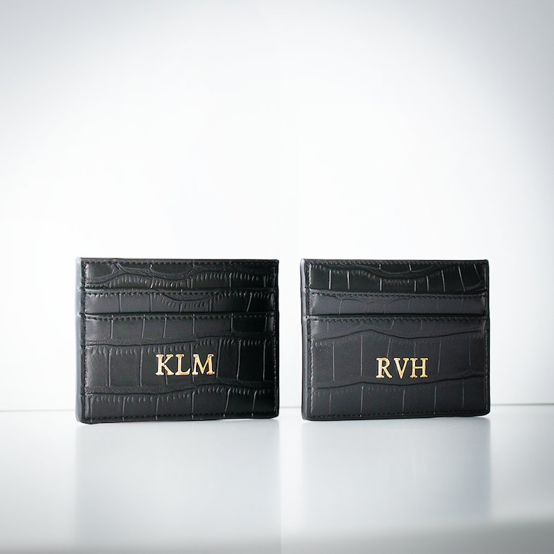 K&ouml;nnte beinhalten: Zwei schwarze Lederkartenhalter mit einem Krokodilpr&auml;gemuster. Die Kartenhalter haben goldene Beschriftungen mit den Initialen "KLM" und "RVH".