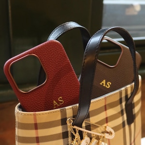 May include: A beige and brown plaid tote bag with black leather handles. Two phone cases are inside the bag, one is red leather with gold initials "AS" and the other is black leather with gold initials "AS". A gold safety pin with a pearl and charm chain dangles from the bag.