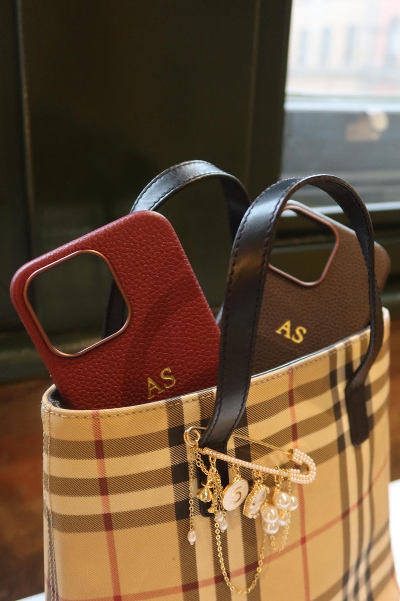 May include: A beige and brown plaid tote bag with black leather handles. Two phone cases are inside the bag, one is red leather with gold initials "AS" and the other is black leather with gold initials "AS". A gold safety pin with a pearl and charm chain dangles from the bag.
