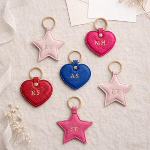 May include: A selection of personalised keyrings in star and heart shapes. The keyrings are in shades of pink, red and blue, with gold-coloured metal rings. Each keyring features embroidered initials.