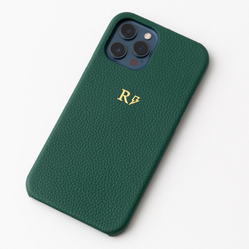 May include: A green leather phone case with a gold monogram "R⚡️" on the back. The case has a textured surface and is designed for a smartphone with a camera bump.