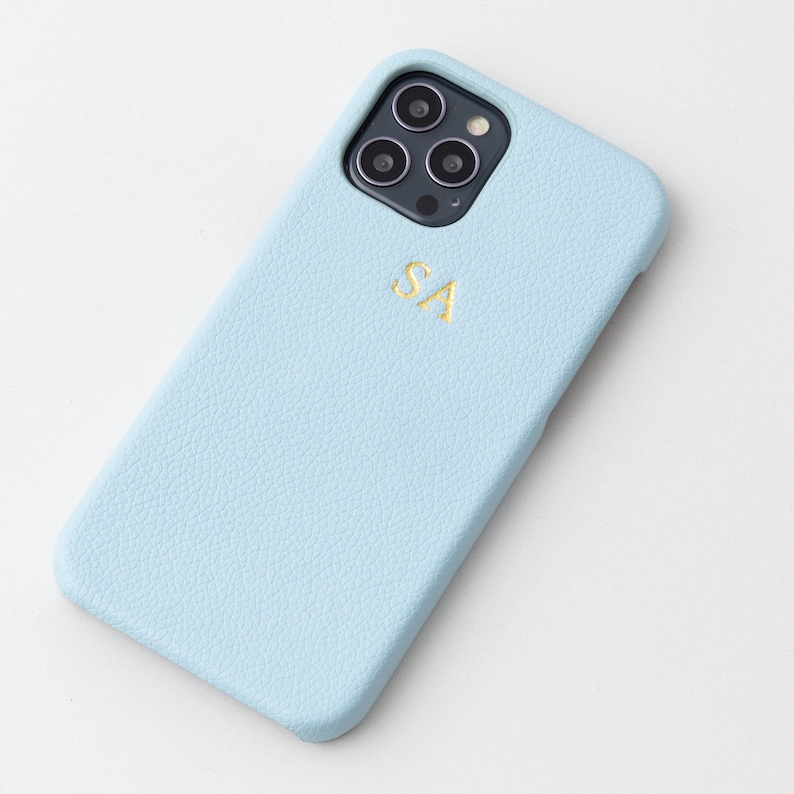 May include: A light blue leather phone case with a gold monogram "SA" and a black camera lens cutout.