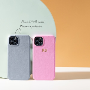 May include: Two iPhone cases, one gray and one pink, with raised edges to protect the camera lens. The pink case has the initials "RS" embossed in gold.