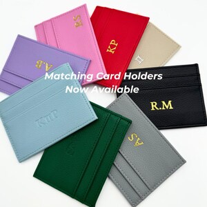 May include: A collection of nine personalized leather card holders in various colors, including black, red, green, blue, gray, pink, and purple. Each card holder has a gold embossed monogram.
