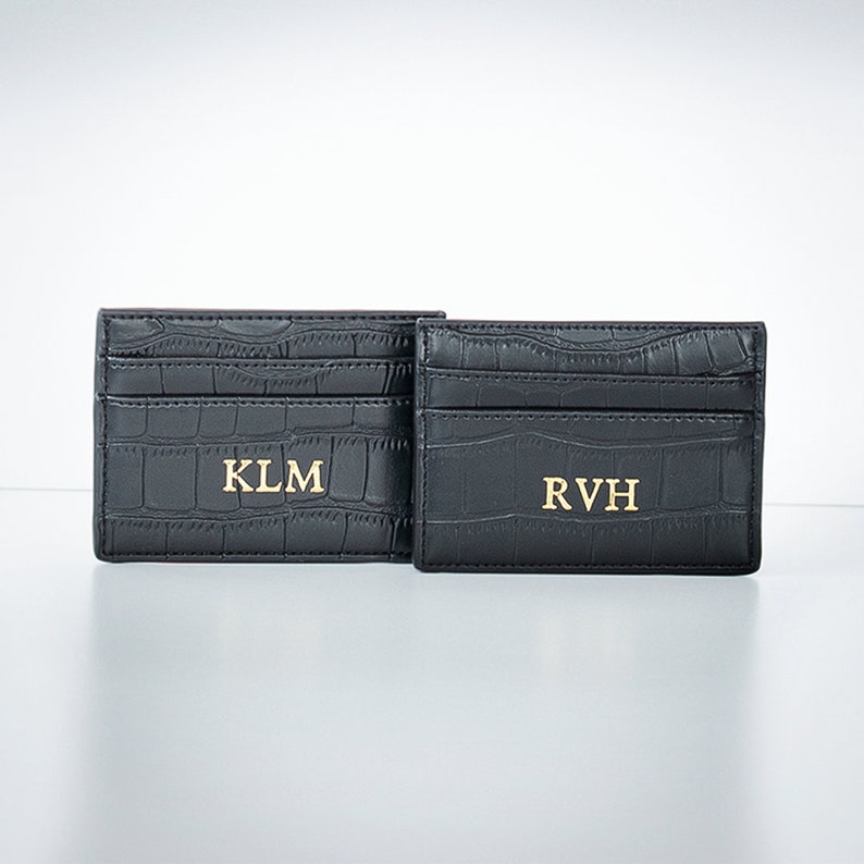 K&ouml;nnte beinhalten: Zwei schwarze Lederkartenhalter mit gepr&auml;gtem Krokodilmuster. Der Kartenhalter links tr&auml;gt die Initialen "KLM" in goldenen Buchstaben. Der Kartenhalter rechts tr&auml;gt die Initialen "RVH" in goldenen Buchstaben.