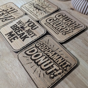 May include: A set of six square wooden coasters with laser-etched text in a comic book style. The phrases include "YOU WILL NOT BREAK ME" and "GODDAMNIT DONUT!" The coasters are light brown.