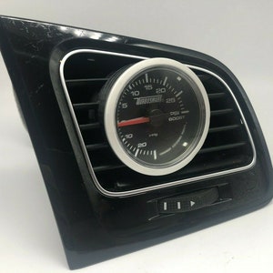 May include: A black car dashboard vent with a silver trim and a white gauge with a black face. The gauge reads "TURBOSMART PSI BOOST" and has a red needle pointing to 10. The gauge is mounted in the vent and has a silver bezel.