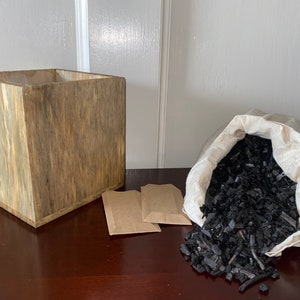 May include: A wooden box with a natural wood grain finish, two brown paper envelopes, and a white canvas bag filled with black charcoal pieces.