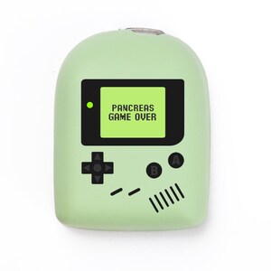 May include: A pale green handheld gaming device with a black screen displaying "PANCREAS GAME OVER." The device has a directional pad, A and B buttons, and speaker details, similar to a retro console. The device is set against a white backdrop.