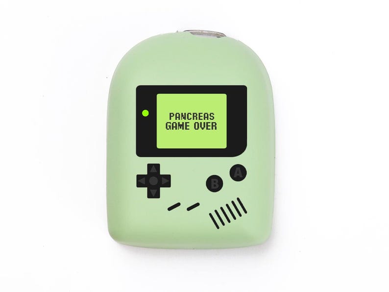 May include: A pale green handheld gaming device with a black screen displaying "PANCREAS GAME OVER." The device has a directional pad, A and B buttons, and speaker details, similar to a retro console. The device is set against a white backdrop.