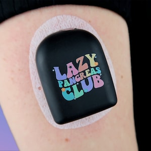 May include: A black medical device with a white adhesive patch attached to a person's arm. The device has a colorful graphic that reads "LAZY PANCREAS CLUB".