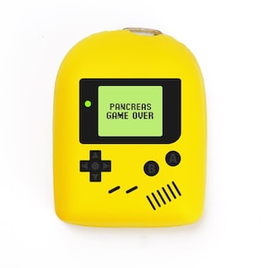 Può includere: Un dispositivo giallo a forma di console di gioco portatile con uno schermo nero che mostra "PANCREAS GAME OVER" in testo verde. Il dispositivo ha pulsanti neri e un D-pad, che assomiglia a un controller di videogiochi classico.