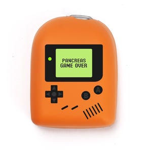May include: An orange novelty item in the shape of a retro handheld console. The screen shows "PANCREAS GAME OVER" in white text on a green background. It has black buttons and a directional pad. A fun, quirky accessory.