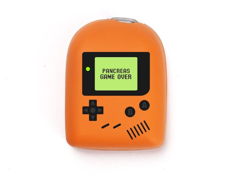 May include: An orange novelty item in the shape of a retro handheld console. The screen shows "PANCREAS GAME OVER" in white text on a green background. It has black buttons and a directional pad. A fun, quirky accessory.