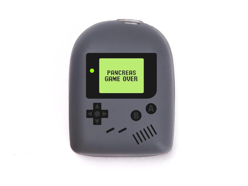 May include: A grey handheld device with a screen displaying "PANCREAS GAME OVER" in white text on a green background. The device has a classic game console design with directional buttons and A/B buttons.