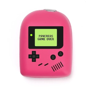 Può includere: Un dispositivo di gioco portatile rosa con uno schermo che mostra "PANCREAS GAME OVER". Il dispositivo presenta un D-pad, pulsanti A e B e prese per altoparlanti. Il design imita una classica console di gioco retrò.