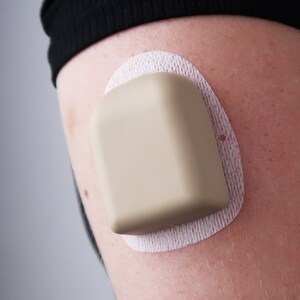 May include: A beige medical device with a white adhesive backing is attached to a person's arm.