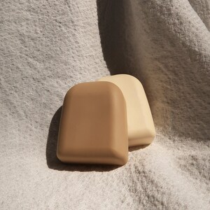 May include: Two small, rectangular-shaped soaps, one beige and one light brown, are sitting on a white, textured fabric.