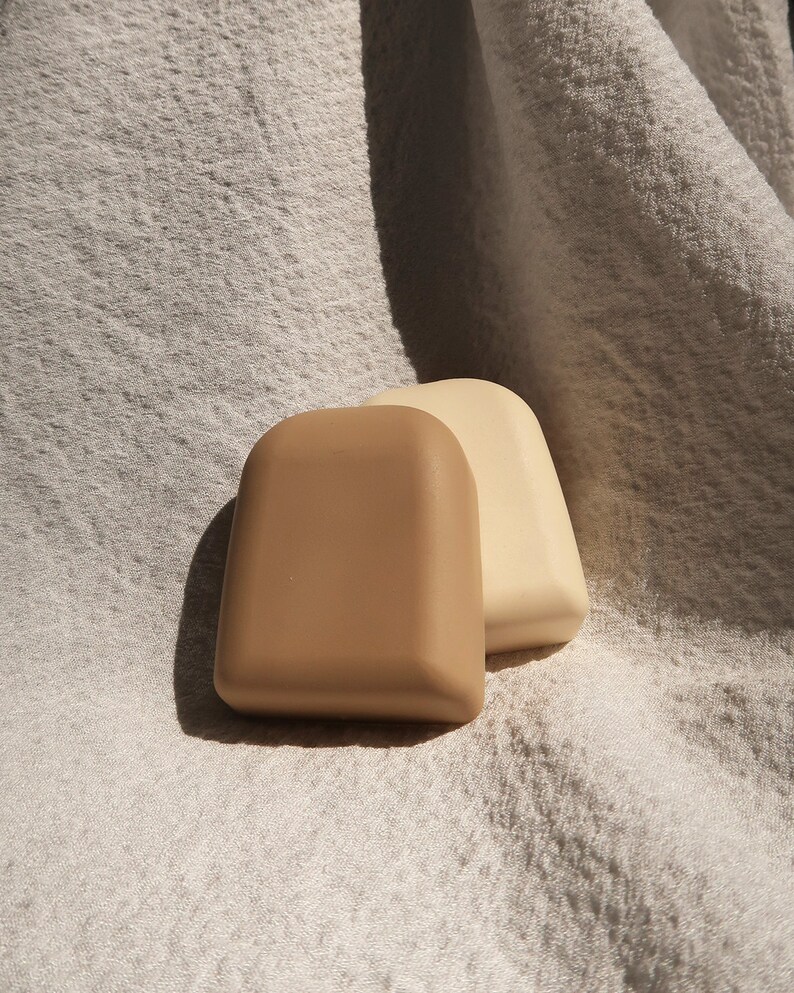 May include: Two small, rectangular-shaped soaps, one beige and one light brown, are sitting on a white, textured fabric.